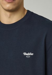 Sweatshirt en coton bleu marine avec un col rond et des manches courtes. Le texte blanc "Buddies" suivi de "Every Sunday" en dessous est brodé sur la poitrine.