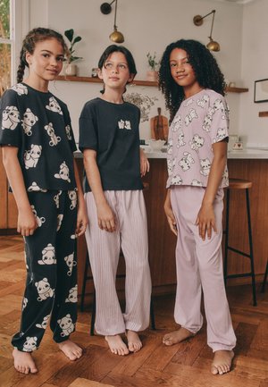 Tre piger står barfodet i et køkken iført nattøj med dyreprint med katte og pandaer i pastel- og mørke farver.