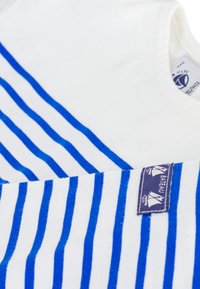 Λευκό μπλουζάκι με οριζόντιες μπλε ρίγες, μαλακό ύφασμα, στρογγυλή λαιμόκοψη και μια μπλε ετικέτα με γραφικά ιστιοπλοΐας ραμμένη στη μία πλευρά.