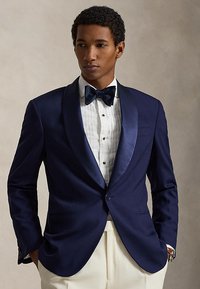 Mann trägt eine marineblaue Smokingjacke, weißes plissiertes Hemd, schwarze Fliege und cremefarbene Hose, die Hände in den Taschen.