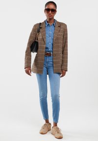 Žena u smeđem kariranom blejzeru, plavoj teksas košulji i trapericama, bež tenisicama, sa sunčanim naočalama i crnom torbom na ramenu.