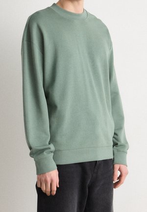 Personne portant un sweat-shirt à manches longues vert sauge avec un petit texte "Hugo" sur la poitrine et un pantalon noir, debout devant un fond uni.