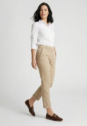 Frau geht, trägt weißes Hemd mit Knöpfen, beige hochgekrempelte Hose und dunkelbraune Loafers mit goldenen Schnallen, vor einfachem hellem Hintergrund.