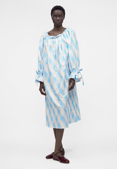 Γυναίκα που φοράει ένα χαλαρό φόρεμα μήκους μέχρι το γόνατο με ανοιχτό μπλε και λευκές διαγώνιες ρίγες, μακριά μανίκια με δεμένες μανσέτες και μπορντό επίπεδα παπούτσια.