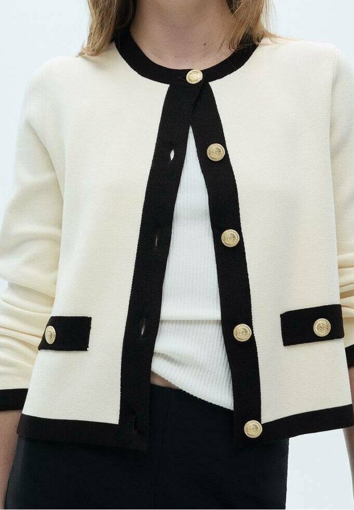 Femme portant un cardigan crème avec des bordures noires et des boutons dorés sur un haut blanc côtelé, associé à un pantalon noir.