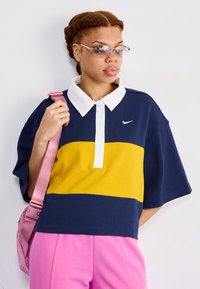 Navy blue and yellow color-blocked polo shirt with a white collar, worn with pink pants; clear-framed sunglasses and a pink shoulder bag.