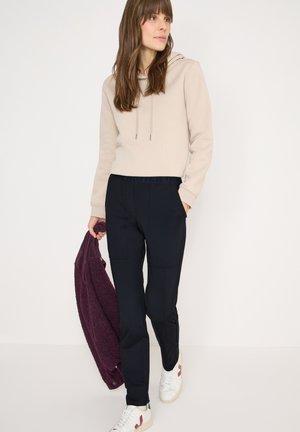 Beige hoodie met trekkoorden, zwarte broek met zakken, eenpaar paars vest vastgehouden, gecombineerd met witte sneakers met bordeauxrode accenten.