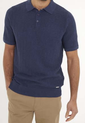 Homme portant un polo bleu marine texturé à manches courtes, rentré dans un pantalon beige, debout avec une main dans la poche sur fond blanc.