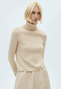 Beige turtleneck sweater lavet af blødt materiale, med en ribbet hals og et kort design, parret med en bølgende beige nederdel.