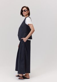 Granatowa kamizelka w paski i szerokie spodnie, w połączeniu z białym t-shirtem. Model ma na sobie czarne okulary przeciwsłoneczne i klapki.