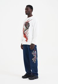 Weißes Langarmhemd mit einem schwarzen und roten Drachenmotiv, kombiniert mit blauen Jeans, die eine bunte Drachenstickerei am Bein haben. Schwarze Sneaker.