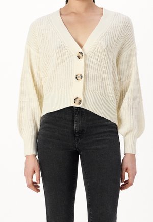 Femme portant un cardigan en maille crème à col en V avec trois boutons en écaille et un jean noir taille haute, mains relaxées le long du corps.
