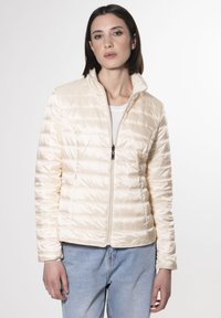 Giacca imbottita color crema con zip frontale, colletto alto, design a righe orizzontali e texture liscia e lucida. Abbinata a jeans azzurri chiari.