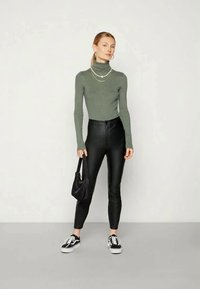 Suéter verde de gola alta com nervuras, combinado com calças de couro pretas de cintura alta e ténis pretos. Inclui uma mala preta e colares em camadas.