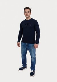 Námornícky dlhý rukáv s čiernym logom USPA, spárovaný s svetlo modrými džínsami a tmavými teniskami. Model stojí na jednoduchom pozadí.