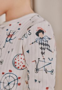 Dziecięca koszula z miękkiej białej tkaniny ma zabawny wzór cyrkowy z klaunami, rowerami i geometrycznymi kształtami w kolorze czerwonym, niebieskim i czarnym.