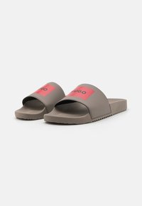 Grijze schuifsandalen hebben een brede band met een rode achtergrond en zwart logo. De gestructureerde voetbed en rubberen zool zorgen voor grip en comfort.