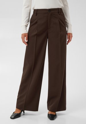 Pantalon marron à jambes larges avec plis sur le devant, porté avec des chaussures noires à talons et un haut blanc à manches longues, visible de la taille aux pieds.