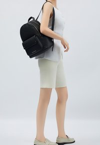 Mustat nahkareput, joissa on teksturoitu pinta, kaksinkertaiset vetoketjut, etutasku ja kultaisenväriset yksityiskohdat. Päälle on puettu vaaleanharmaa paita ja kremväriset shortsit.