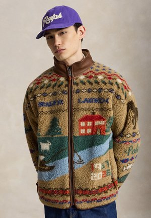 Jeune homme portant une veste polaire colorée avec des motifs nature et cabane et une casquette violette avec l'inscription "Ralph".