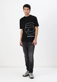 Black cotton t-shirt featuring a white outline graphic of sunglasses and face, with "KARL LAGERFELD" text, paired with distressed dark jeans and sneakers.