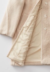 Cappotto in pile beige con tessuto esterno testurizzato, quattro bottoni bianchi e una fodera beige liscia, dotato di una tasca laterale esterna.