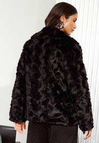 Cappotto in pelliccia sintetica nera con una superficie testurizzata e soffice. Presenta un ampio colletto e maniche lunghe. Abbinato a pantaloni a pois.