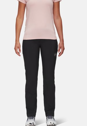 Mujer vistiendo una camiseta de manga corta color rosa claro, pantalones ajustados negros con un logo en el muslo y zapatillas deportivas blancas con detalles en gris y negro.