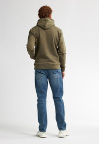 Felpa verde oliva con cappuccio regolabile, tessuto strutturato e polsini a costine. Indossata con jeans blu e sneakers bianche.