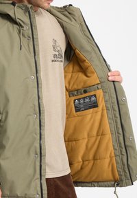 Veste isolante vert olive avec une doublure en velours jaune moutarde. Comprend une fermeture éclair, des boutons-pression et une étiquette dans la poche intérieure.