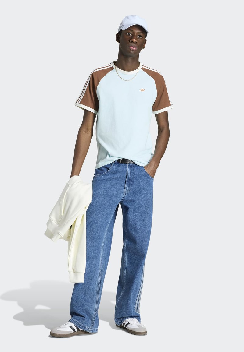 Junger Mann trägt eine weiße Kappe, hellblaues und braunes T-Shirt, blaue weite Jeans und weiße Turnschuhe, hält einen weißen Sweatshirt in einer Hand.