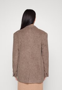 Hnědý oversize blazer z vlněné směsi s klopovým límcem, polstrovanými rameny a jemnou texturou. Obsahuje jeden rozventilovaný otvor a dlouhé rukávy.