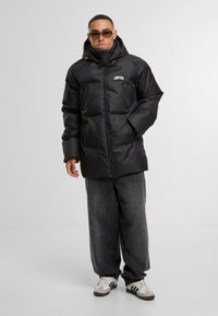 Svart puffjakke med hette, med front glidelås, "UNFAIR" logo, og to sidelommer. Bært over brede grå denim bukser og sko.