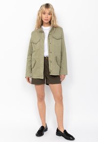 Giacca verde oliva con bottoni a pressione, due tasche sul petto e un taglio dritto, indossata sopra una t-shirt bianca e pantaloni corti marroni con una vestibilità classica.