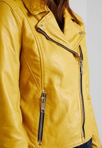 Giacca da motociclista in pelle gialla con design aderente, cerniere argentate, colletto a pressione e tasche laterali. Texture liscia con dettagli di cucitura sottili.