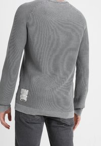 Graues Langarmshirt mit geribbelter Textur, Rundhalsausschnitt und einer strukturierten Applikation unten links. Das Shirt hat einen lässigen Schnitt und kontrastierende Nähte.