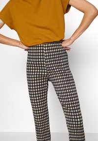 Pantalon à carreaux noirs et beiges taille haute avec un design plissé, associé à un haut à manches courtes couleur moutarde. Texture lisse et coupe ajustée.