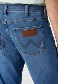 Dark blue denim jeans with a straight cut, featuring a brown leather patch, decorative stitching, and two back pockets with a distinctive design.