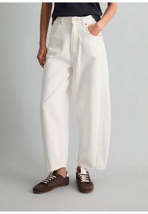 Osoba nosząca luźne białe spodnie z wysokim stanem i brązowe trampki, z jedną ręką w kieszeni, stojąca na jednolitym szarym tle.