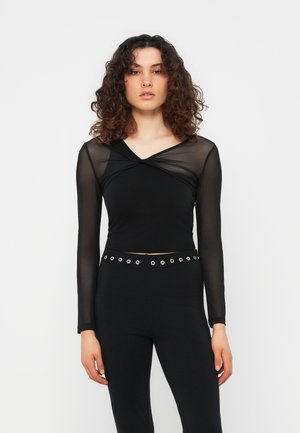 Γυναίκα με σγουρά μαλλιά, που φοράει μαύρο μακρυμάνικο τοπ από δίχτυ και μαύρο παντελόνι με ασημί ζώνη με τρουκ, στέκεται μπροστά σε λευκό φόντο.