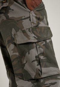 Cargo pants in camouflage pattern, featuring multi-pocket design, gray and green tones, with sturdy fabric and visible stitching details.