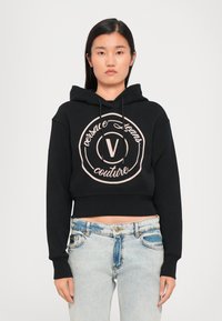 Juodas trumpas džemperis su platinta jose ir apvaliu rožiniu logotipu, ant kurio yra tekstas „versace jeans couture“. Derinamas su šviesiai mėlynomis džinsomis.