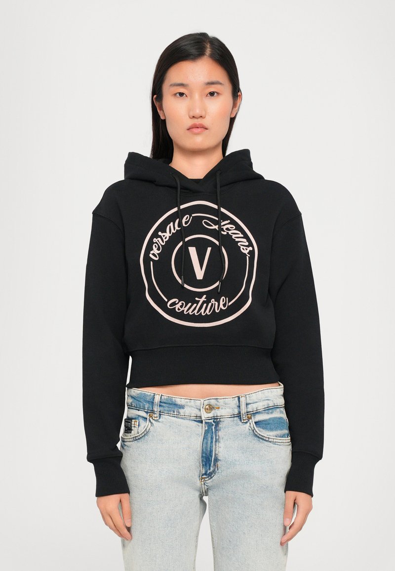 Juodas trumpas džemperis su platinta jose ir apvaliu rožiniu logotipu, ant kurio yra tekstas „versace jeans couture“. Derinamas su šviesiai mėlynomis džinsomis.