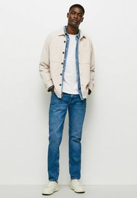 Jachetă de corduroy bej deschis peste o cămașă de denim albastră, un tricou alb, blugi albaștri și adidași albi. Croială slim cu detalii cu nasturi.