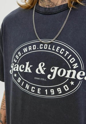 Donker marineblauw t-shirt met ronde hals en een wit grafisch logo met "Jack & Jones" en "Sinds 1990." Zichtbare zilveren ketting.