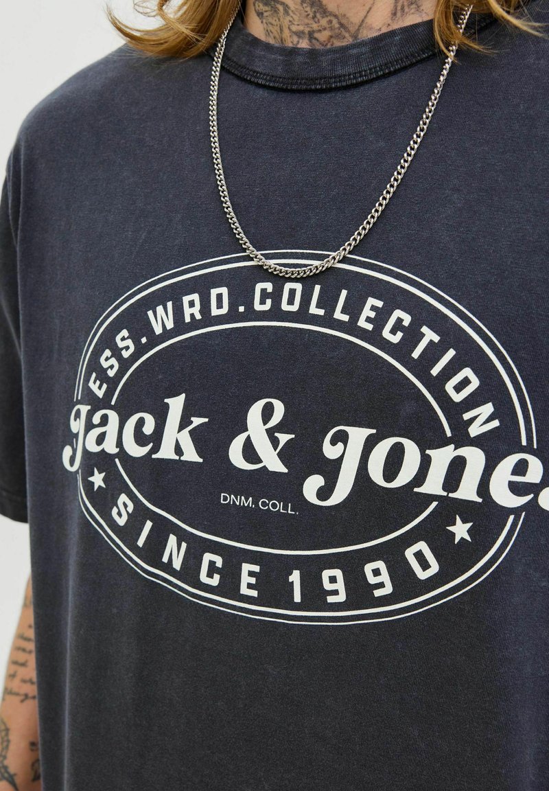 Tmavomodré tričko s okrúhlym výstrihom s bielym grafickým logom "Jack & Jones" a "Since 1990." Viditeľný strieborný reťazový náhrdelník.