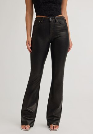 Femme portant un pantalon en cuir noir taille haute évasé avec des poches avant, associé à des sandales beige à bouts ouverts et un top court noir.