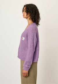 Paarse, fluffy cardigan met een diepe V-hals, lange mouwen en een voorzak. Draag over een textuur, olijfgroene kledingstuk.