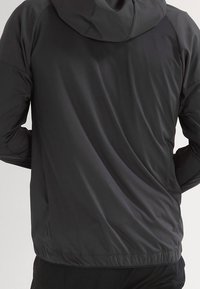 Svart lättviktsjacka med huva, slät textur, elastiska ärmslut och beskuren nederkant. Bakifrån visar den en minimalistisk design och materialets glans.