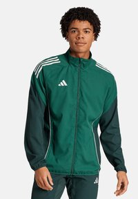 Groene sportjack met contrasterende donkergroene mouwen, witte strepen op de schouders, rits aan de voorkant en elastische manchetten. Bevat het Adidas-logo.